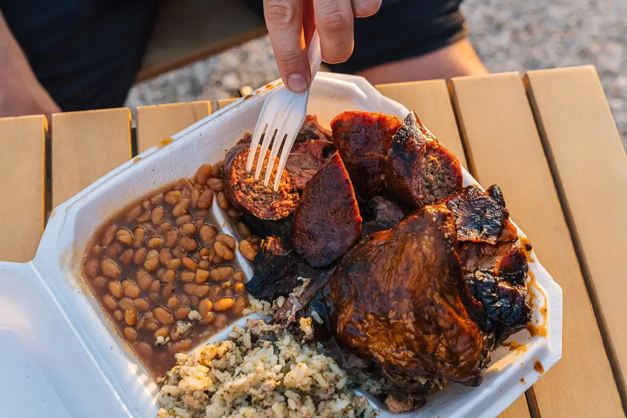 Person using a fork to take a piece of Smoked Sausage from a Takeout Container full of Smoked BBQ Beans, Chicken Thighs & Drumsticks, and Dirty Rice.