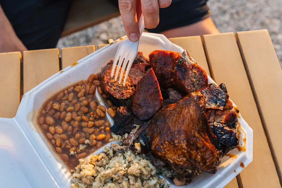 Person using a fork to take a piece of Smoked Sausage from a Takeout Container full of Smoked BBQ Beans, Chicken Thighs & Drumsticks, and Dirty Rice.